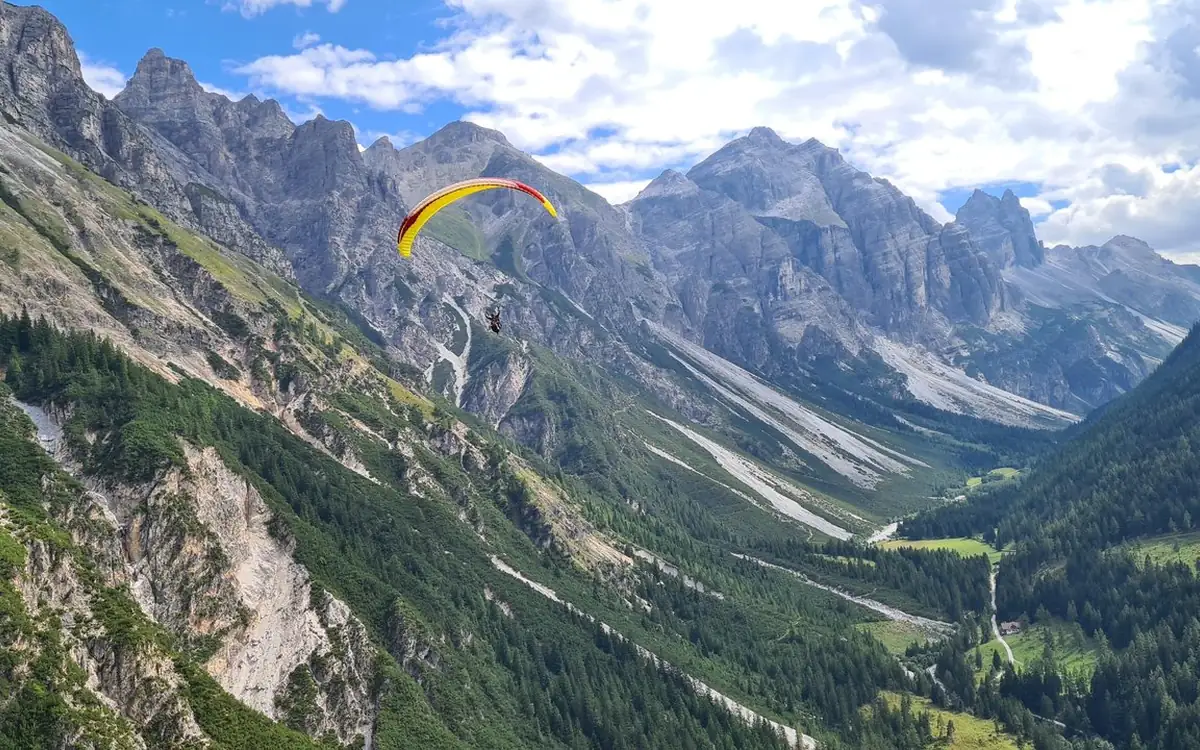 A yellow and red paraglider over a wooded slope below grey limestone peaks