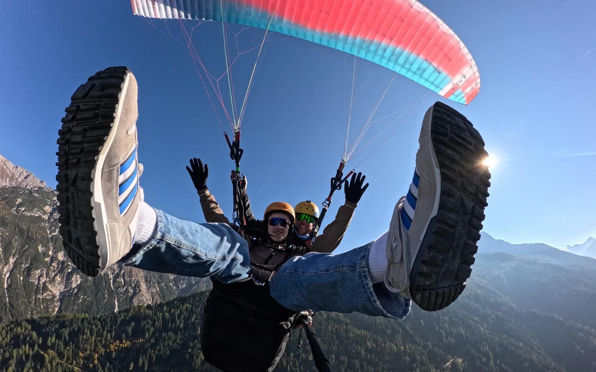 Selfie from a tandem paragliding flight: a passenger's feet raised into frame, both people in helmets under a red and turquoise wing, dark forest far below