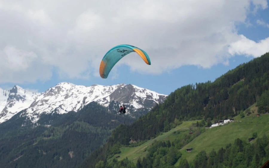 A teal and orange paraglider wing above snow-topped peaks and a green wooded hillside