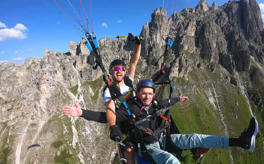 Two flyers under one wing passing a ridge of grey rock spires above green slopes