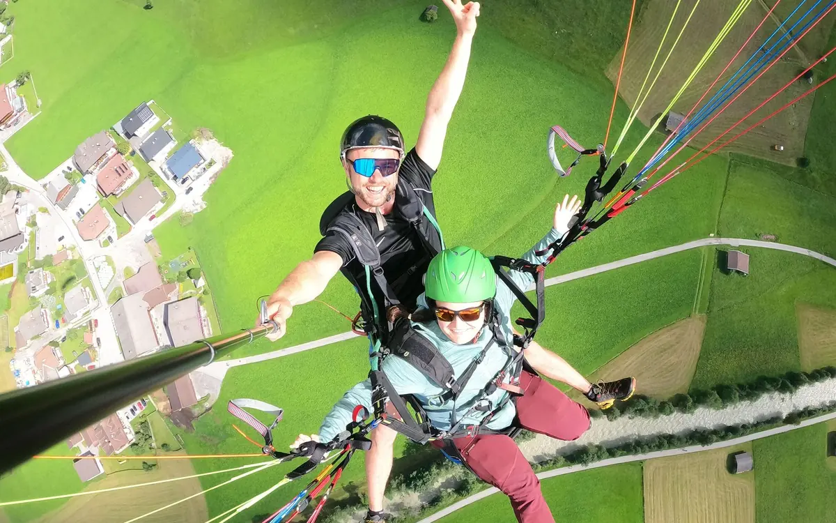 Looking down from a tandem paragliding flight over the Stubai Valley: two people under one wing, lines fanning out above them, mown green meadows and village roofs far below